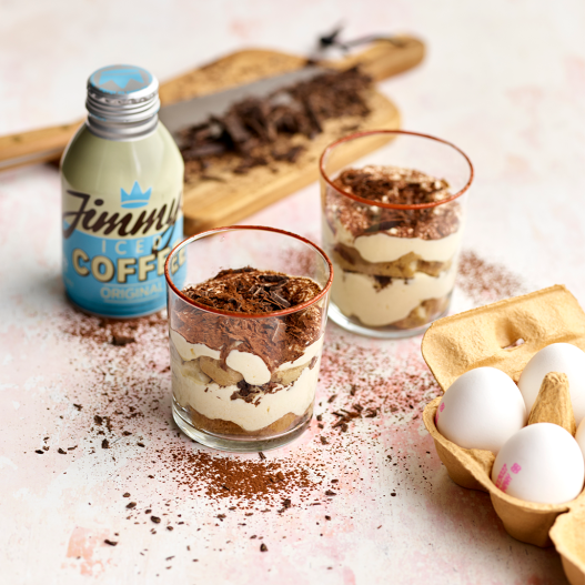 2 tiramisu desserts against a pale pink background, surrounded by a box of Respectful eggs with white shells and a light blue bottle of Jimmy's iced coffee. In the background is a wooden chopping board containing chopped chocolate.