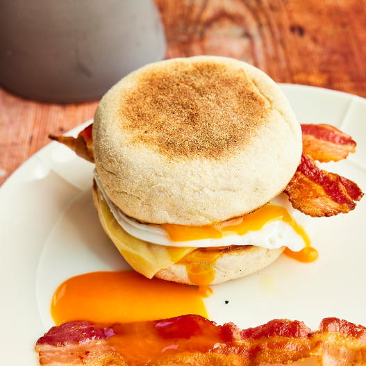 A hand holds a Respectful eggs breakfast sandwich, an English muffin with fried egg and streaky bacon