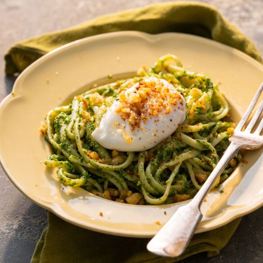 Kale pesto pasta with a poached egg sitting on top, all sprinkled with crispy breadcrumbs. This meal sits in a cream bowl which has a silver fork resting on the side.