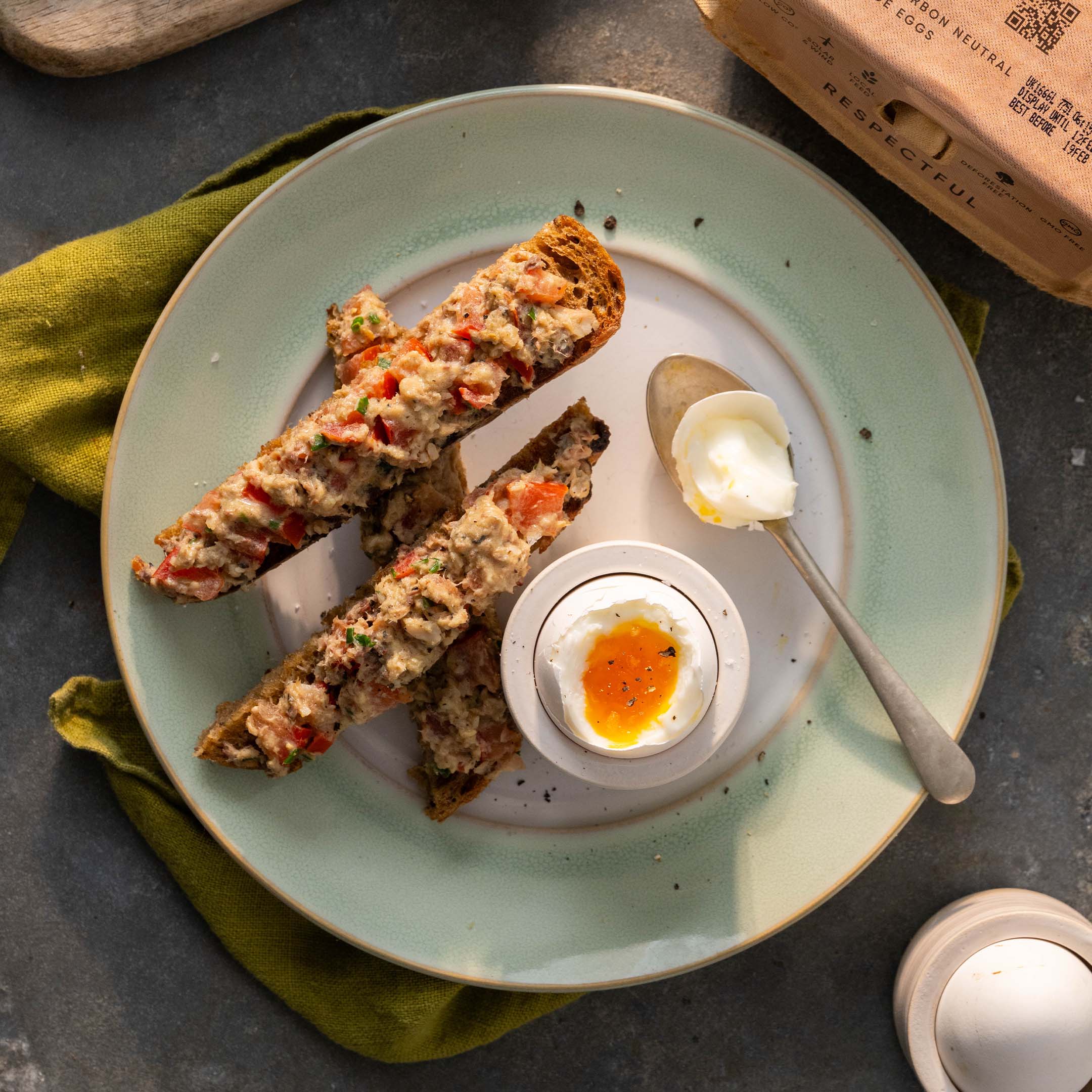 A boiled egg sits in an egg cup with the top of it cut off, exposing the orange yolk inside. Next to the boiled egg lies three devilled sardine soldiers - three lengths of toast with mashed sardines on top.