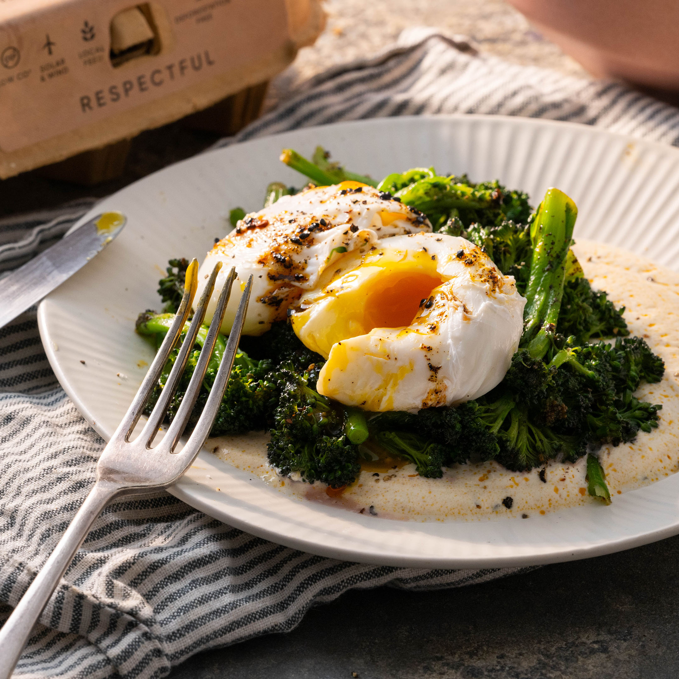 This poached egg, sitting on top of broccoli and spiced eggs is sitting on a ribbed white plate. One of the two poached eggs has been popped and the yolk is running into the broccoli below.