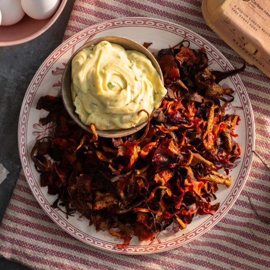 A red rimmed plate filled with homemade vegetable crisps with a small bowl of black pepper and chive mayonnaise nestled in the side. The crisps are made up of potato, beetroot, carrot and parsnip skins that have been air fried to make them crispy.