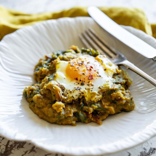 Beautifully cooked lentil Dahl with a baked egg on top sits on a white plate shaped like a flower