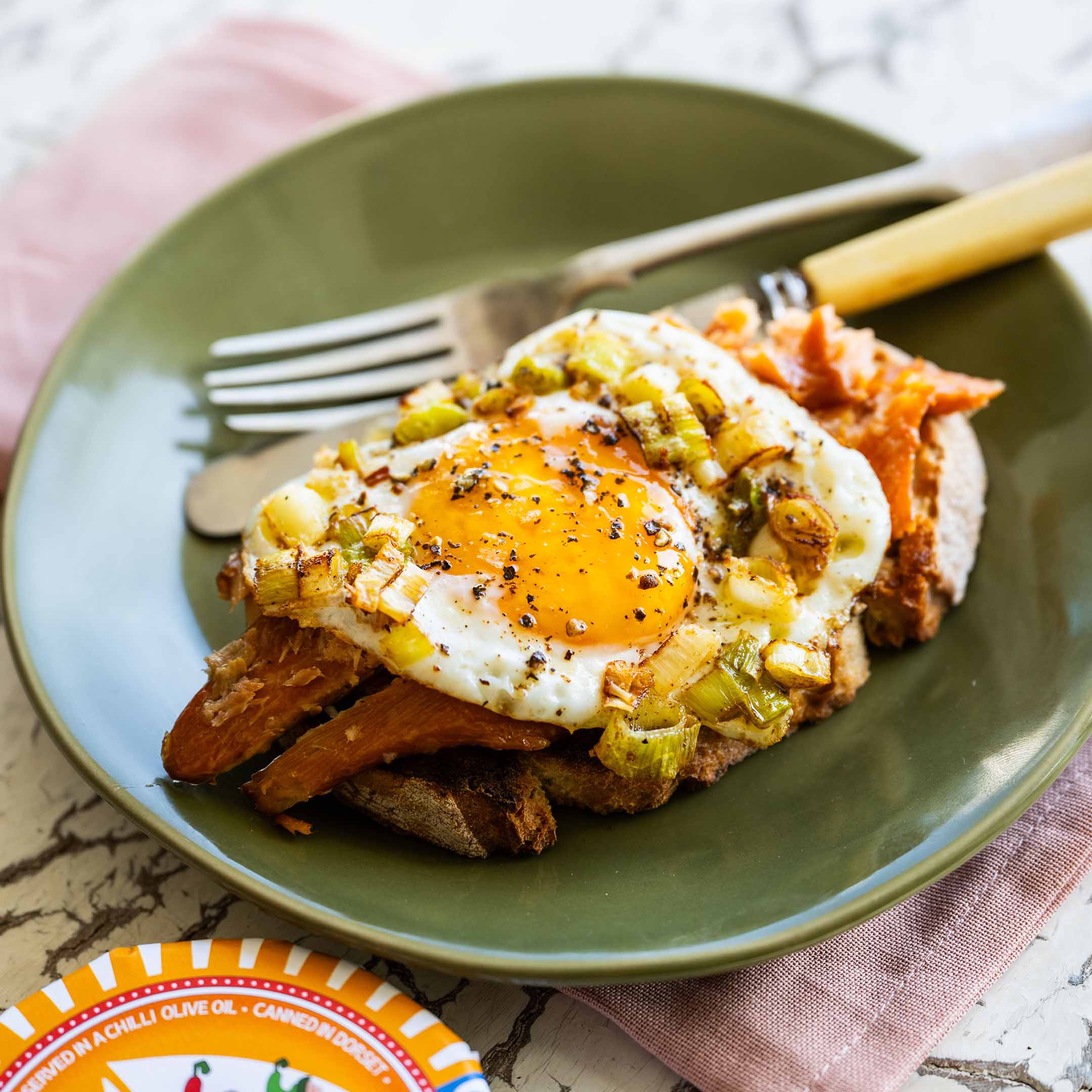 A green plate sits next to a Sea Sisters tin of rainbow trout in chilli oil. On the plate is a round of sourdough toast, with the rainbow trout flaked on top, and a fried egg crowns off the entire meal.
